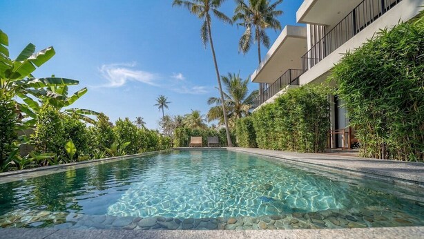 Ein klares Schwimmbecken mit Steinrand, umgeben von üppiger Vegetation und Palmen, mit einem modernen Gebäude an einer Seite.