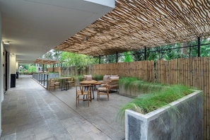 Een overdekt terras met tafels en stoelen, een bamboescherm en een plantenbak met gras.