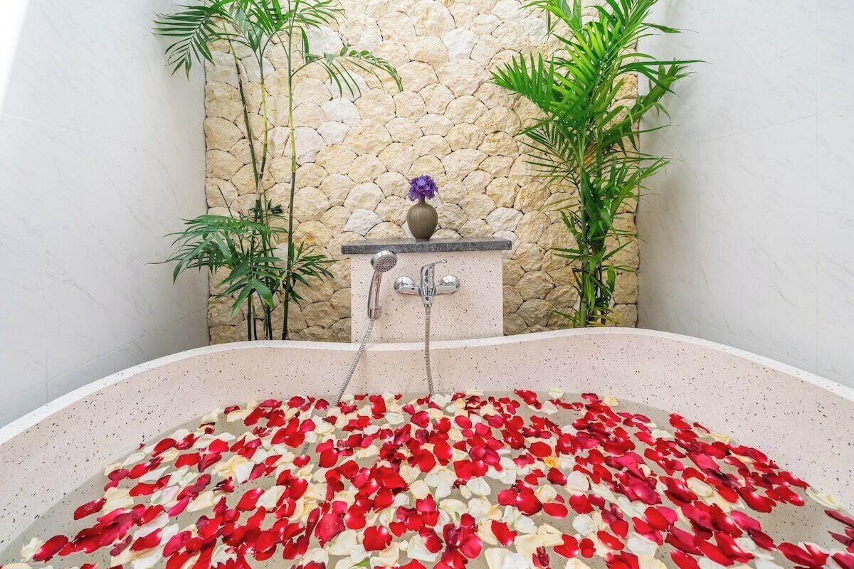 Una bañera llena de pétalos de rosa, una pared de piedra y plantas de bambú.