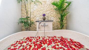 Una bañera llena de pétalos de rosa, una pared de piedra y plantas de bambú.