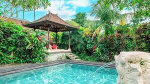 Una piscina con una escultura de piedra, rodeada de exuberante vegetación y un pabellón tradicional.