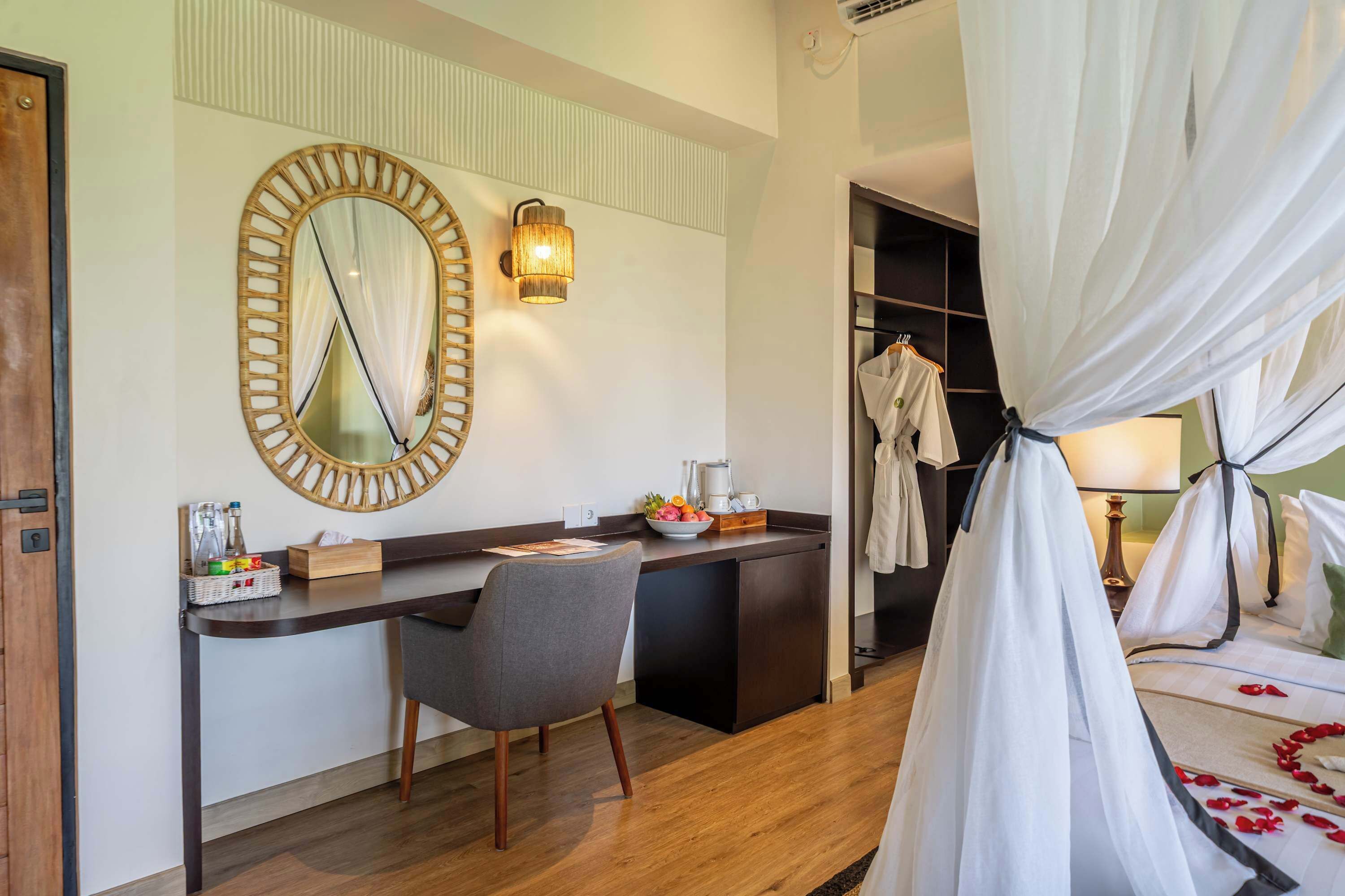 A hotel room with a bed, a desk with a chair, a mirror, a wall-mounted lamp, and a wardrobe.