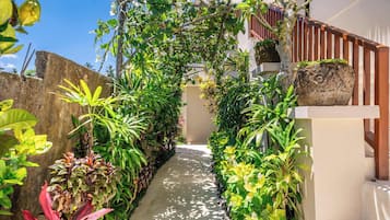 Un sentier étroit bordé d’une végétation luxuriante et d’une clôture en bois mène à un bâtiment dont le toit est recouvert de chaume.