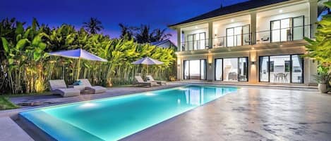 A modern two-story house with a swimming pool, outdoor seating, and lush tropical vegetation.