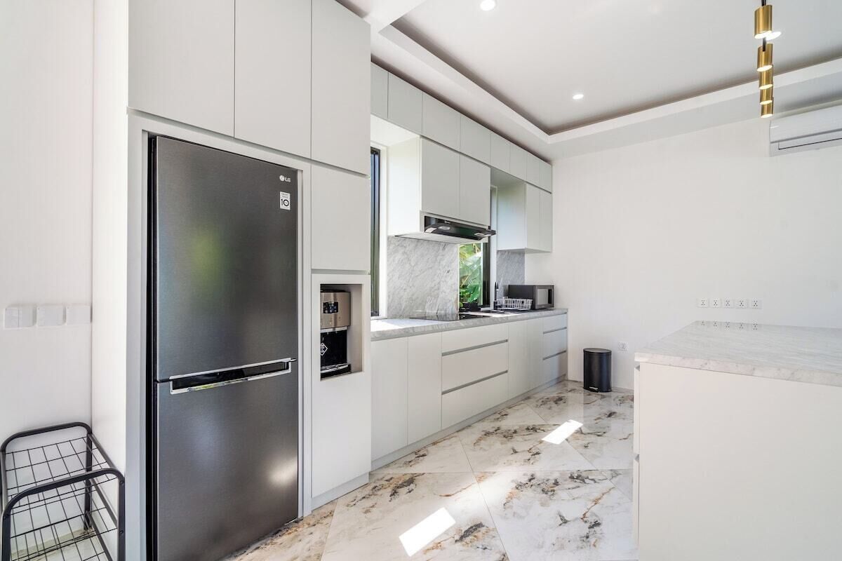 A modern kitchen with a stainless steel refrigerator, white cabinetry, a marble countertop, and a black trash bin.