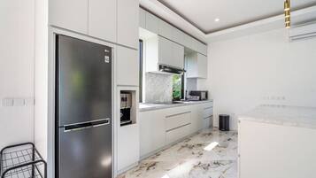 A modern kitchen with a stainless steel refrigerator, white cabinetry, a marble countertop, and a black trash bin.