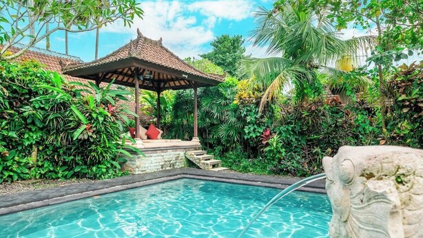 Una piscina con una escultura de piedra, rodeada de exuberante vegetación y un pabellón tradicional.