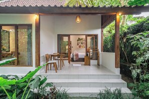 Una habitación con cama, una mesa y sillas, una puerta que da a otra estancia, y vista a un jardín.