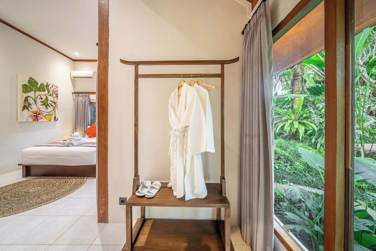 A bedroom with a bed, a wooden bench with robes and slippers, and a view of greenery through a window.