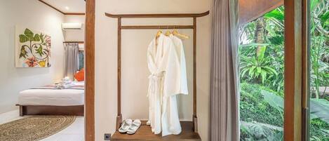 A bedroom with a bed, a wooden bench with robes and slippers, and a view of greenery through a window.