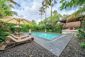 Una piscina con sillas de descanso y una sombrilla, rodeada de vegetación tropical y una estructura de piedra.