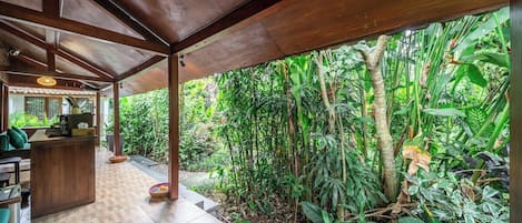 A covered outdoor area with a wooden floor, surrounded by lush tropical vegetation.