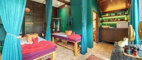 A room with two beds, a massage table, a mirror, and a shelf with bottles.