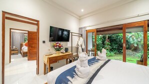 A hotel room with a bed, a desk, a television, and a view of greenery outside. - Elegant New 2BR in a Complex w Spa, 2 Pools+Garden (Ubud)