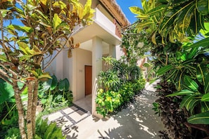 Un sentier bordé de végétation luxuriante traverse un jardin tropical et mène à un bâtiment doté d’une porte en bois, le tout baigné par un ciel dégagé.