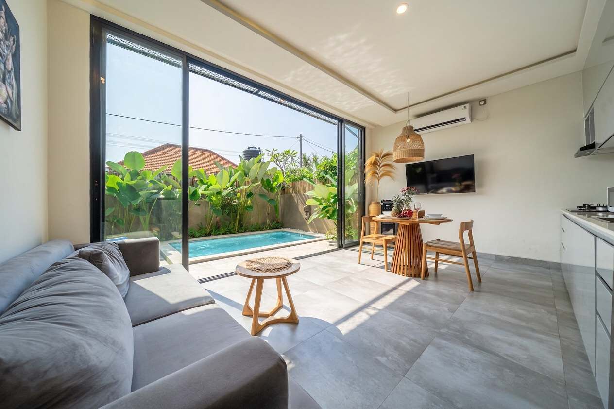 Ein modernes Wohnzimmer mit einer großen Schiebeglastür, die zu einem Pool führt, einem Essbereich mit Tisch und Stühlen sowie einer Küche mit Schränken und einem Fernseher.