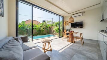 Ein modernes Wohnzimmer mit einer großen Schiebeglastür, die zu einem Pool führt, einem Essbereich mit Tisch und Stühlen sowie einer Küche mit Schränken und einem Fernseher.