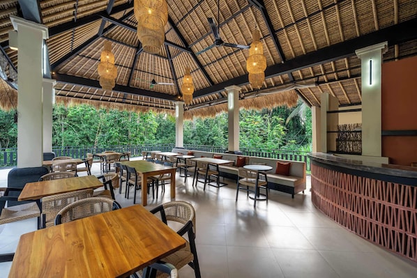 An outdoor seating area with tables and chairs, a thatched roof, and a view of greenery.