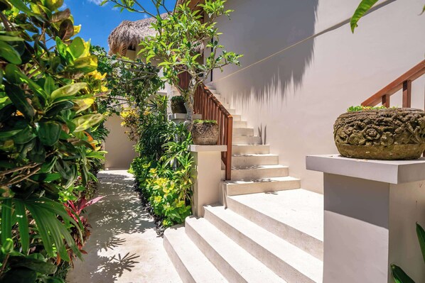 A staircase leading up to a building with a thatched roof, surrounded by lush greenery and a clear sky.