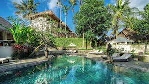 A clear swimming pool with lounge chairs and a statue, surrounded by tropical vegetation and a building with a thatched roof.