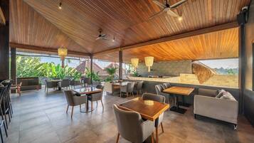 A spacious indoor area with wooden ceiling, multiple seating arrangements including tables and chairs, and a view of the outside.