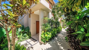A tropical garden pathway leading to a building with a wooden door, surrounded by lush greenery and a clear sky. - Chic Ricefield View Suite in Mesari Hotel Ubud (Gianyar Regency)