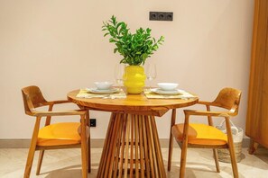 A dining area with a round wooden table, two chairs with orange cushions, a yellow vase with green foliage, and white plates and glasses. - Stylish Flat with Kitchen in Peaceful Part of Ubud (Gianyar Regency)