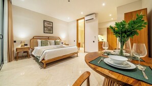 A hotel room with a bed, bedside table, lamp, and a dining area with a table set for two. - Cozy 1BR in Serene Ubud - Kitchen, Balcony, Pool (Gianyar Regency)