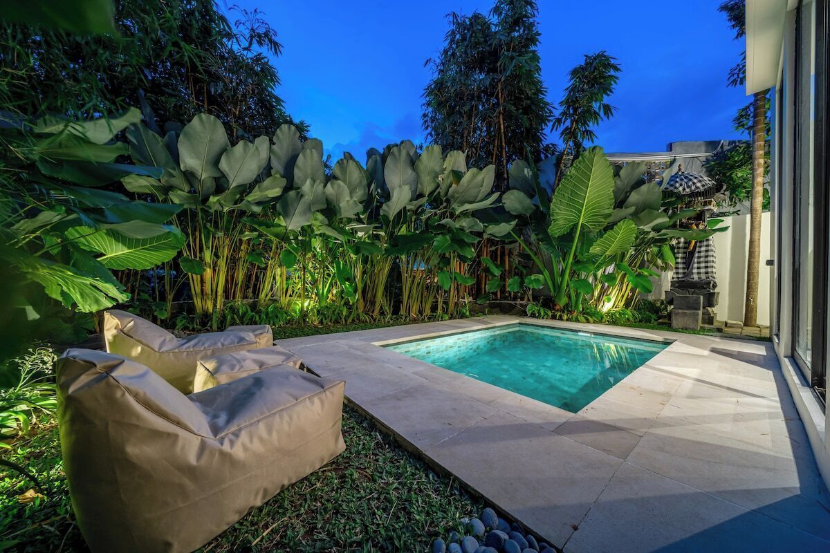 Ein Swimmingpool mit umlaufender Terrasse, einer Liege und üppiger tropischer Vegetation.