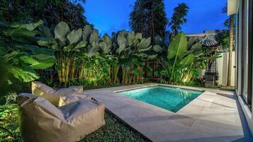 Ein Swimmingpool mit umlaufender Terrasse, einer Liege und üppiger tropischer Vegetation.
