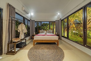 Un dormitorio con una cama grande, una mesita de noche, un perchero y vistas a la vegetación a través de amplias ventanas.