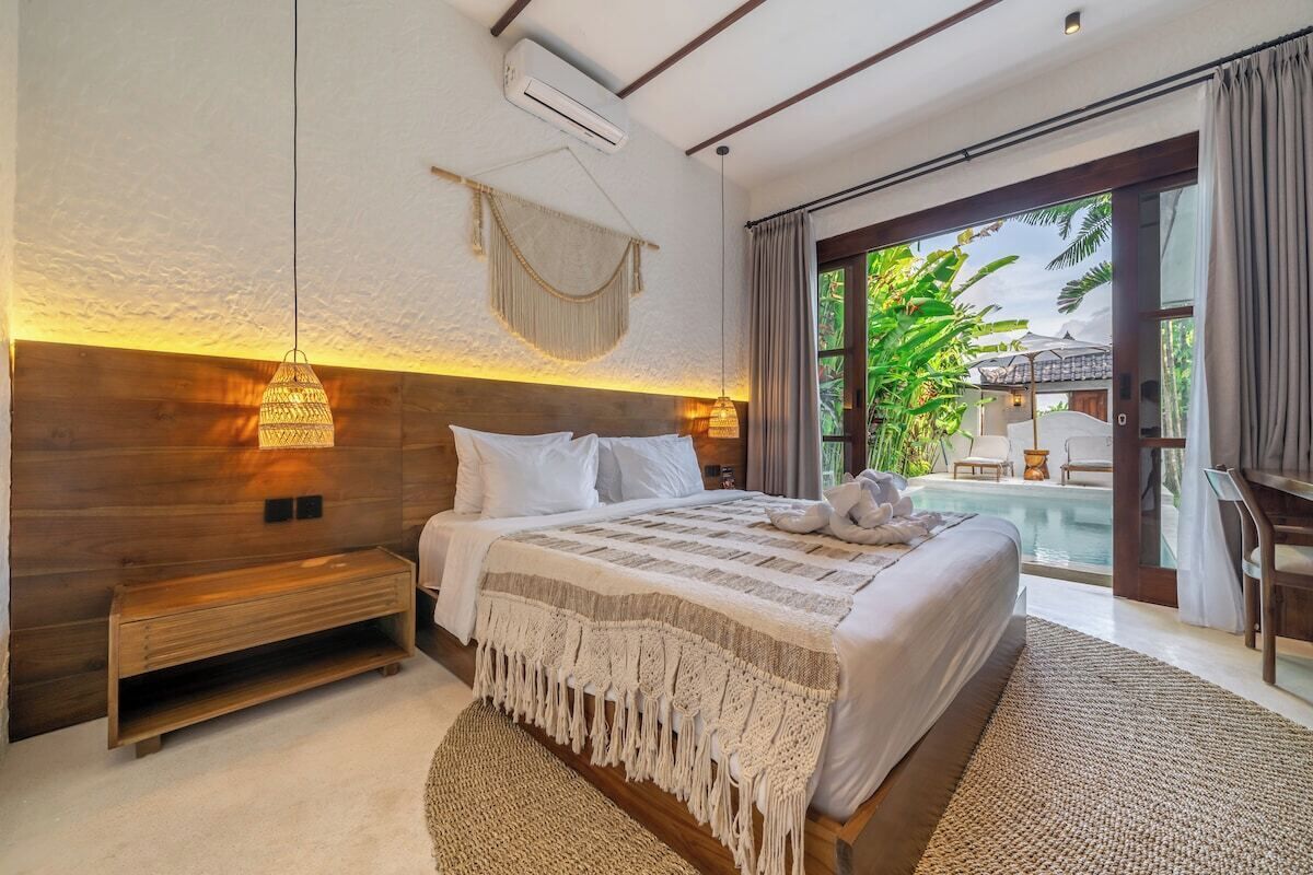 A bedroom with a large bed, a wooden headboard, a bedside table, a hanging light fixture, and a view of an outdoor pool area.