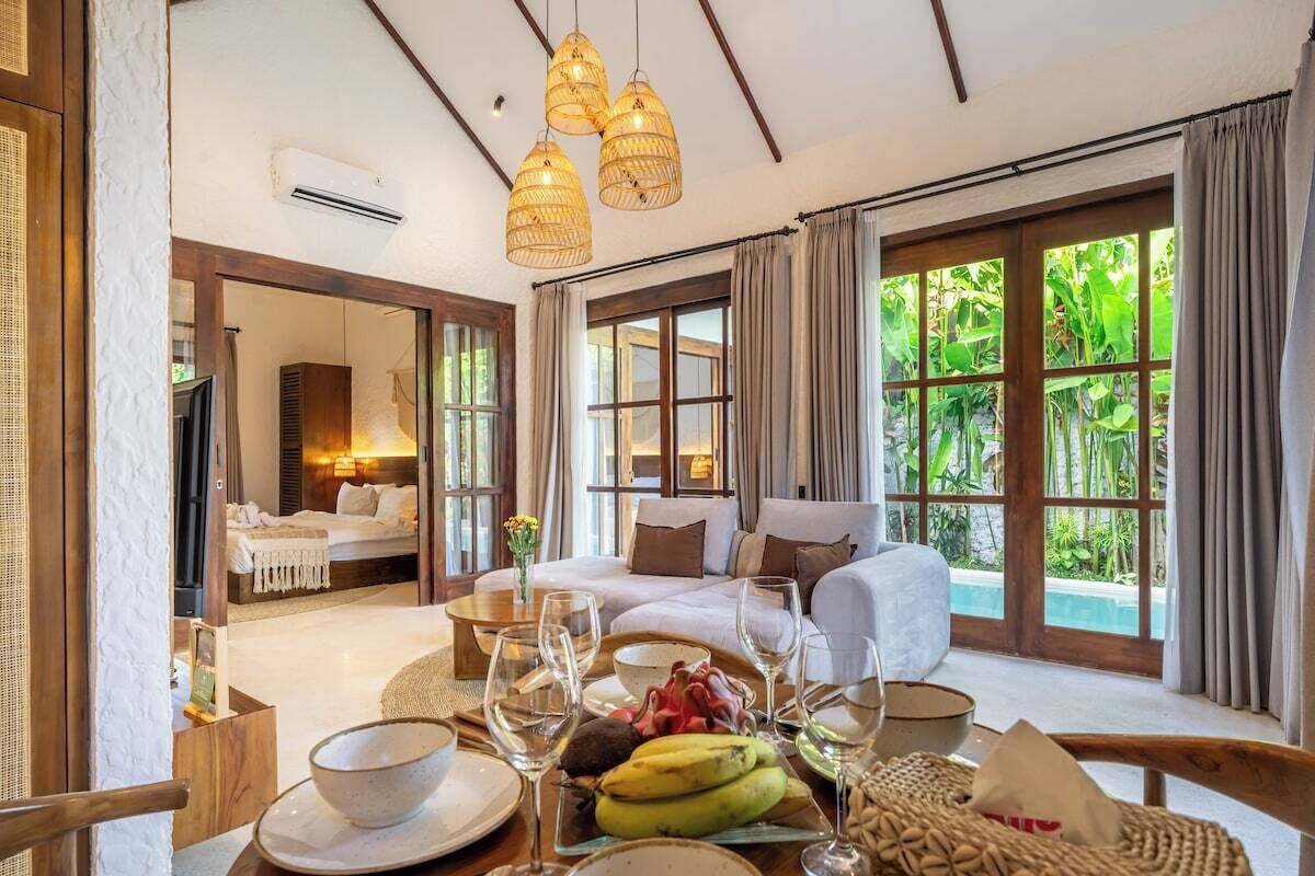 A room with a bed, a sofa, a dining table with glasses and fruits, and large windows with curtains.
