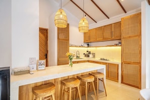 A kitchen with wooden cabinets, a central island with stools, hanging wicker light fixtures, and a microwave. - Designer 3BR Villa w Garden Bath & Pool • Ubud (Gianyar Regency)