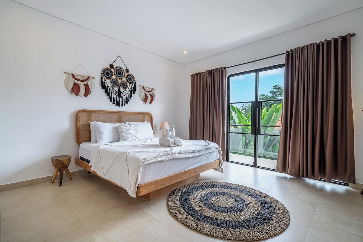 Een slaapkamer met een groot bed, een rond geweven vloerkleed en uitzicht op groen via een schuifdeur.