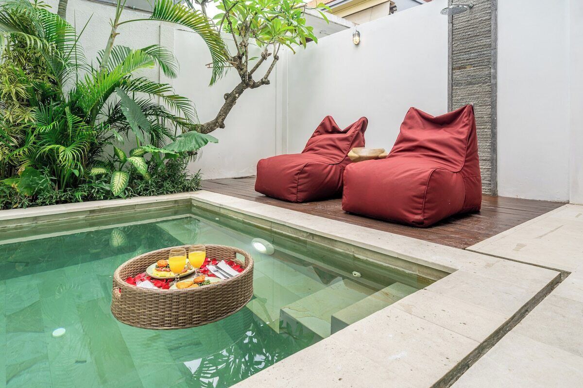 A pool area with a wicker basket containing food and drinks, surrounded by red cushions and lush greenery.