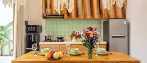 A modern kitchen with a dining table set for two, featuring a fruit bowl, a vase of flowers, and a refrigerator.