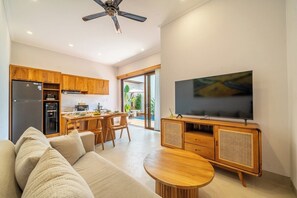 A modern living room with a sofa, a dining area with chairs, a kitchen with wooden cabinets, a television, and a ceiling fan. - Cassandra 3 - Styish 5BR in 500 m to Sanur Beach w Pool+Balcony (Kota Denpasar)