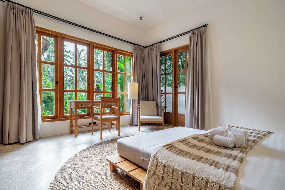 A bedroom with a bed, a desk, and a chair near large windows.
