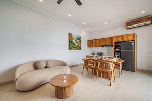 A modern living room with a sofa, dining table, and chairs. There is a kitchen area with cabinets and appliances. - Chic 3BR in Prime Sanur w Pool & Beach Access (Kota Denpasar)