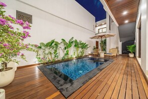 A modern outdoor pool area with wooden decking, surrounded by potted plants and a seating area with a table and chairs. - Chic 3BR in Prime Sanur w Pool & Beach Access (Kota Denpasar)
