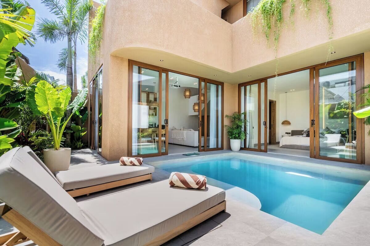 A modern pool area with lounge chairs, a glass-enclosed living space, and tropical plants.