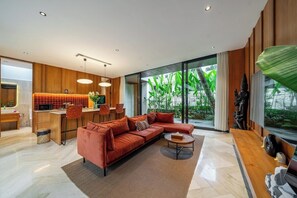 A modern living room with a sectional sofa, a coffee table, and a view of greenery through large windows. - X7 Ubud - Tropical Ubud's 3BR Gem w Pool & Outdoor Dining (Gianyar Regency)