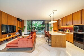 Een moderne keuken met een centraal eiland, een eethoek met barkrukken, een grote flatscreen-televisie en een gezellige zithoek met een bank.