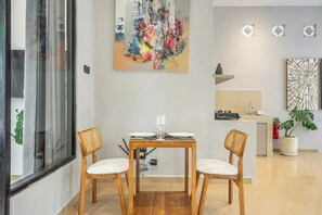 A dining area with a table set for two, a modern abstract painting above, and a kitchen in the background. - Serene 2BR Villa in Ubud Complex w Spa & 2 Pools (Kabupaten Gianyar)
