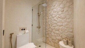 Un baño con ducha envuelta en vidrio, inodoro blanco y pared de piedra.
