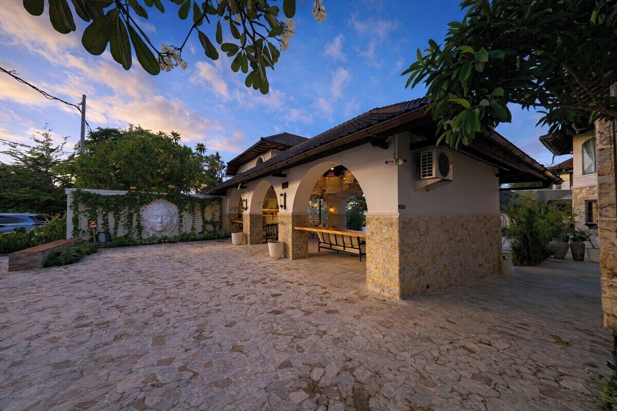 A stone-paved courtyard with a covered outdoor seating area, a stone wall with a carved face, and a clear sky.