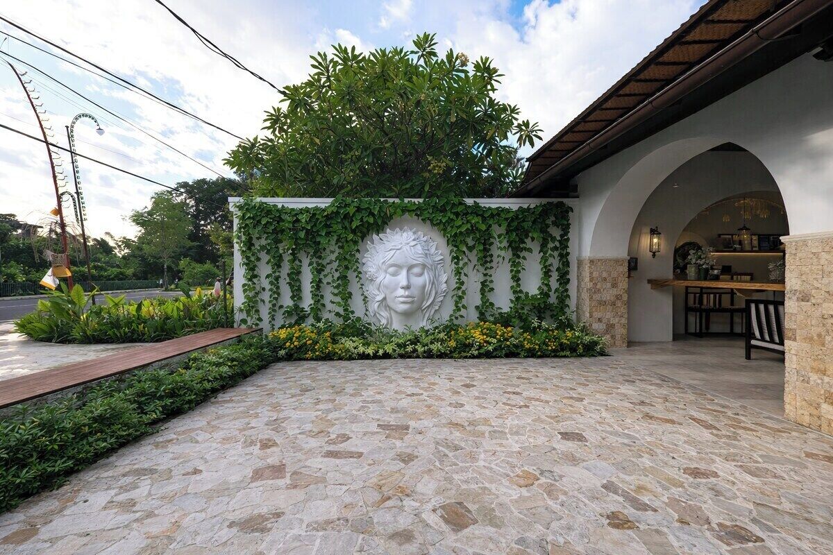 Une entrée dallée de pierres, avec un mur décoratif orné d’un visage sculpté, entouré de verdure et d’une structure en arc.