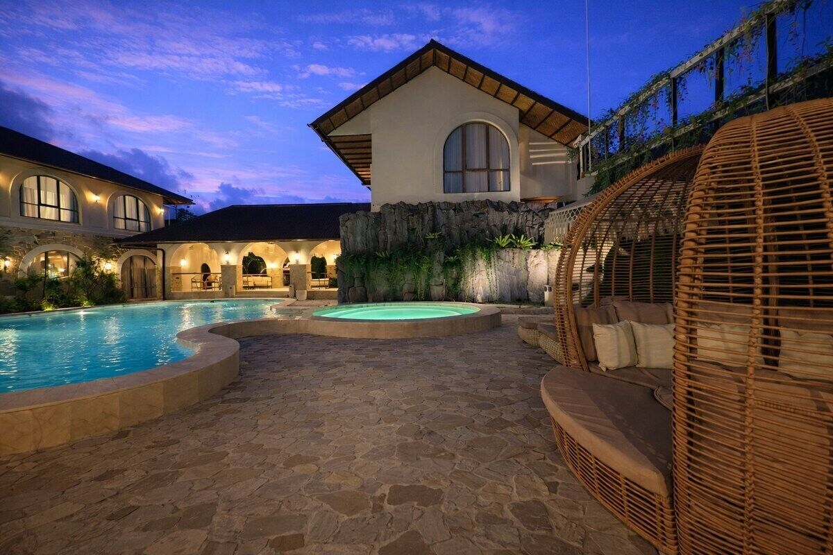 A pool area with a stone floor, a lounge chair, and a building with a large window and arched doorways.