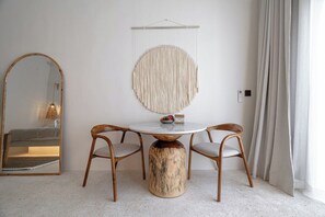 Quarto com uma mesa redonda, duas cadeiras, um detalhe decorativo em macramé, um espelho e uma janela com cortinas.